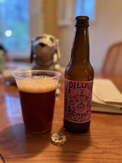 A plastic cup of deep ruby red beer next to a brown bottle with a pink label