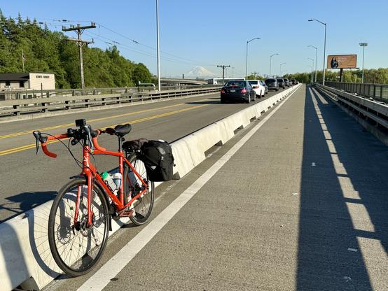 My orange acoustic bicycle leans against a concrete mini jersey barrier in a future 2-way protected bike lane on a bridge over railroad tracks with a view of a distant mountain
