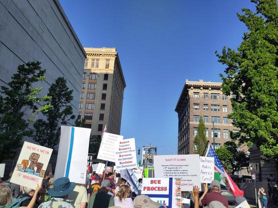 Photo of a march in  progress, with most marchers carrying signs with anti-Trump, pro-democracy slogans. It is a sunny May afternoon in downtown Tacoma, and the march is heading east down 11th Street toward the Murray Morgan Bridge. The destination is the extremely evil for-profit Northwest Detention Center, an ICE processing facility owned by GEO Group.