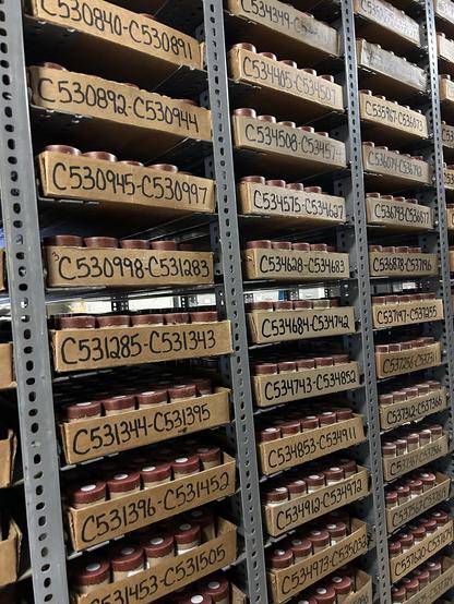 A storage area with metal shelves displaying numerous cardboard boxes, each containing small round containers. The boxes are labeled with alphanumeric codes.