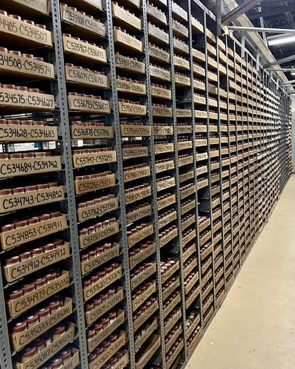 A large storage area filled with metal shelving units containing numerous boxes labeled with serial numbers. The boxes are organized in rows, creating a systematic and orderly appearance. The floor is concrete, and the lighting is bright.