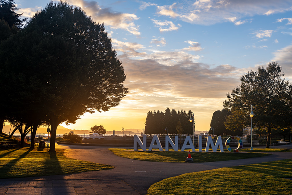 An oceanfront park at sunrise. Large block letters spell out the name "Nanaimo", with most letters in white except the "o", which is painted in a series of solid colours. The sky is slightly cloudy, divided into orange for sunrise and blue toward the top of the photo. Some trees border the image in silhouette. A lamppost still has its lamp light lit.