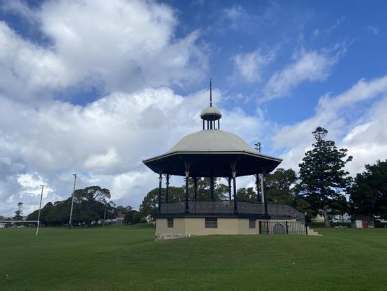 Historic band rotunda set in lush green parkland with talk trees in the background. Round white metal roof with a mini round roof on top and spike. Lots of wrought iron lace railings and decorative detail on the columns. Big white fluffy clouds but blue sky making it through.