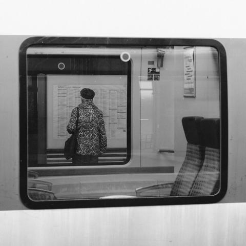 Das Schwarz-Weiß-Foto zeigt durch die Fenster eine Zuges eine Frau, die sich einen Abfahrtsplan ansieht. Im Vordergrund ist der Innenraum eines Zuges mit einigen leeren Sitzen. Die Frau steht mit dem Rücken zum Betrachter. Ihre Jacke hat ein zweifarbiges Muster. Das Muster erinnert an ein Labyrinth – wie auch der Abfahrtsplan.

The black and white photo shows a woman looking at a departure schedule through the windows of a train. In the foreground is the interior of a train with some empty seats. The woman is standing with her back to the viewer. Her jacket has a two-tone pattern. The pattern is reminiscent of a labyrinth - as is the timetable.