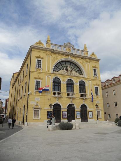 The image showcases a historic yellow building featuring ornate architectural details and large windows. It displays flags and is situated in a plaza with people walking. The structure is identified as the Croatian National Theatre in Split.