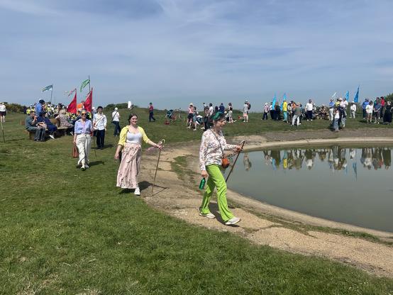 Woman in green trousers and young woman in long pink skirt bith holding staffs walking around Warren Hill Dew Pond, a round, man made lake (for sheep) in the south downs. Other people and flags in background