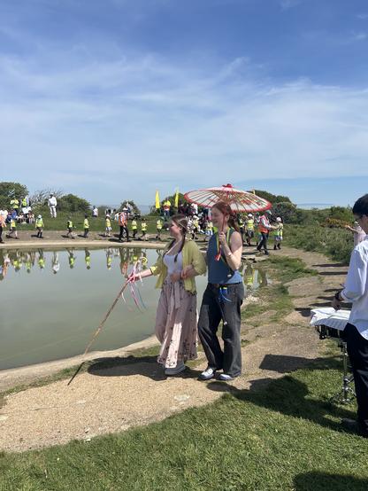 Too young women walking round a round lake, more people in background, blue sky. You can see it is very hot. One of the youbg women holding a nice paper umbrella.