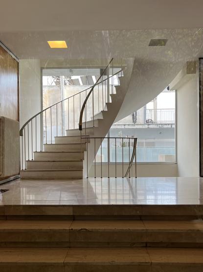 The entrance of an apartment building from perhaps the 1960s. A minimalist staircase swoops up from a marble foyer towards the upper floors and doglegs down to the basement. There’s wood paneling on the left hand wall, unornamented, with a white painted radiator, and stairs from street level to the foyer. The really striking feature is that the rear wall of the foyer is plate glass, flooding the space with light and highlighting the curve of the suspended stairs. Through the glass, the balconies and flat roofs of other buildings, solar water heaters.