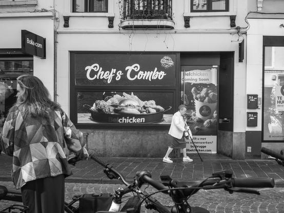 The black and white image depicts a street scene in Bruges, Belgium. In the foreground, a woman with long hair, wearing a patterned jacket, stands next to a bicycle. She is facing away from the camera, looking at a storefront. A woman with a cane is walking on the sidewalk, wearing a white jacket and shorts, moving towards the right side of the image.
The storefront features a large advertisement for "Chef's Combo" with an image of a dish containing chicken. Below the main advertisement, there is a smaller sign that reads "Belchicken Coming Soon."