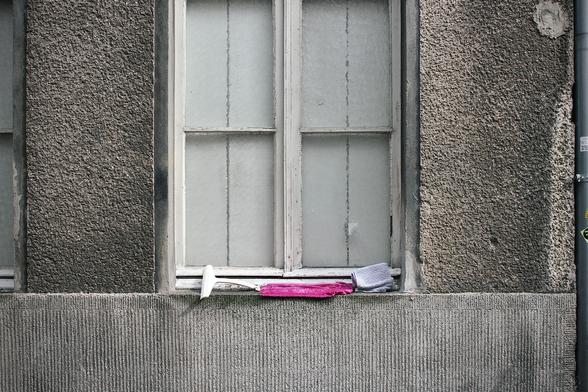 ein fenster in einer alten, grauen hauswand. das fenster ist alt und aus holz. der weiße lack blättert langsam ab. oben ist das fenster angeschnitten und nicht ganz auf dem bild.
auf dem fenstersims liegen ein abzieher und zwei putztücher, eines rosa, eines grau. der putz der wand ist rau und uneben. hinter den strukturierten fensterscheiben sind weiße fensterläden aus holz. der gesamte eindruck ist schlicht und etwas verfallen.