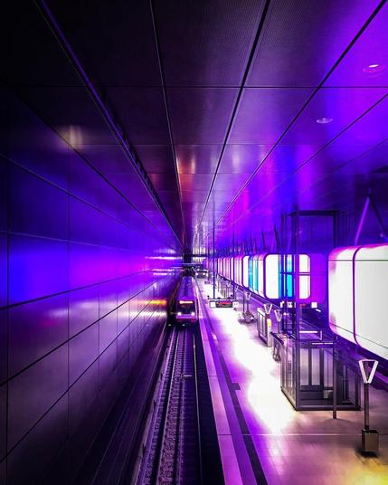A modern subway station in Hamburg’s Hafencity, illuminated by striking purple and blue light panels. A train approaches the platform, surrounded by sleek architecture and glowing reflections.