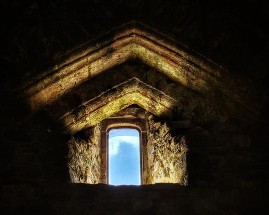 A small, arched window set into a dark, rough stone wall, allowing bright light to stream in.
