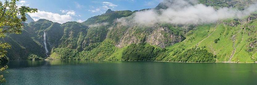 Vue panoramique du lac d'Oô, entouré de montagnes verdoyantes et baigné par une lumière douce. Situé au sud de la Gascogne, ce lac alpin offre un spectacle naturel captivant, caractérisé par ses eaux cristallines et les cascades qui l'alimentent en arrière-plan. L'environnement évoque calme et sérénité.