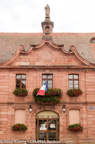 The image depicts the façade of a "HOTEL DE VILLE," which is a town hall in somewhere in France. The building is constructed from pinkish-red stone, typical of the region, and features a symmetrical design with a central clock tower topped by a statue. The clock face is black with gold numerals and hands, and the tower is adorned with a coat of arms above the clock. The building has two windows on the upper floor, each with a flower box filled with red and pink flowers. The French flag is prominently displayed in the center window. The entrance is marked by an arched doorway with a glass door, flanked by lantern-style lights. The roof is covered with red tiles, and the sky is overcast.