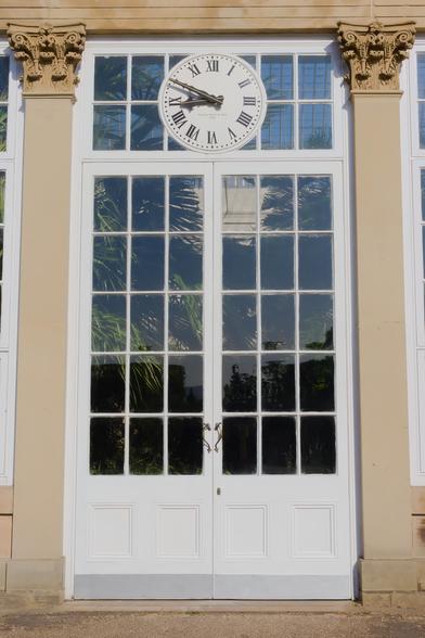 The picture represents a tall, elegant white-painted wooden and glass-paneled door sits at the center of a neoclassical greenhouse façade at the Sheffield Botanical Gardens. Above the door is a round clock with Roman numerals, framed by large, symmetrical Corinthian-style pilasters topped with ornate capitals. The clock face reads “William Potts & Sons, Leeds,” nodding to a respected historic clockmaker. The glass panes reflect palm fronds and the structure's roof, hinting at the lush environment within the glasshouse. The entire composition evokes a refined 19th-century style, blending function, beauty, and botanical wonder.
