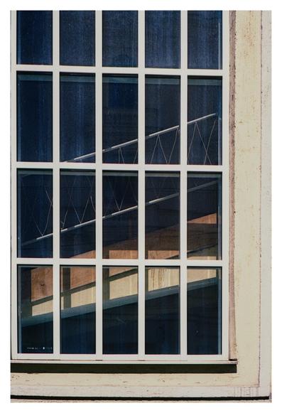 Color photograph of a window of a former factory in Vienna. Through the large window you can see a wooden ramp or staircase with a metal railing. The whole picture is in beige, as the beige-yellow façade of the factory building can be seen to the right and below the window, which corresponds wonderfully with the wood inside.