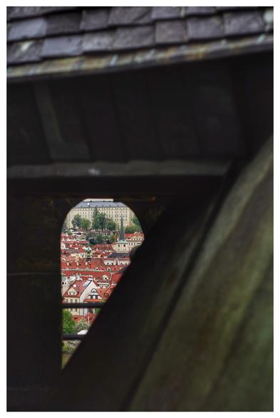 Hochformatiges Farbfoto mit weißem Rand. Blick durch ein Gewirr aus dunklen Dächern. Ganz hinten ist ein kleines, oben abgerundetes Fenster, durch das man viele Gebäude mit roten Dächern sieht.