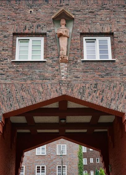 Bâtiment d’entrée sobre en briques dont on voit deux fenêtres et une sculpture stylisée représentant une femme et à ses pieds un enfant, portant l’année gravée 1927. Le bas du bâtiment permet de circuler à pied ou en voiture, on voit d’autres bâtiments en briques par l’ouverture.