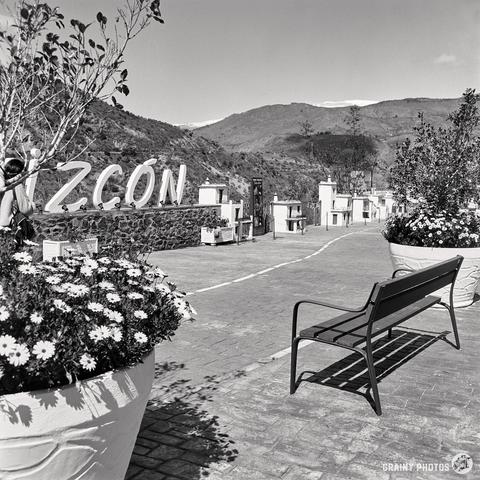 A black and white photograph captures a brick pathway in Torvizcón, Spain, lined with large flower pots and scale models of village houses. A bench stands on the RHS. In the background, you can see a mountain landscape, while a partially visible sign of the village name stands out against the background