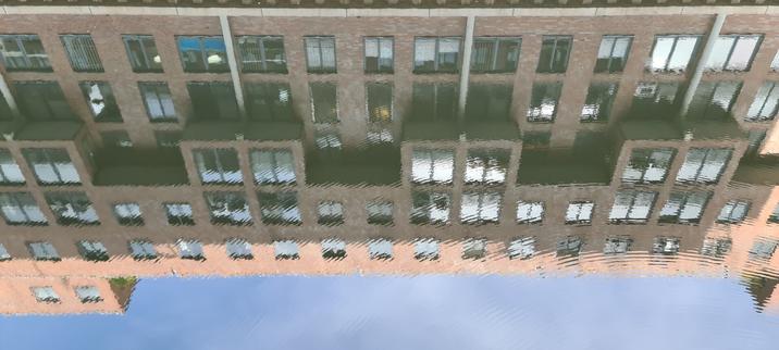 Rotes Backsteingebäude mit Flachdach und vielen Fenstern spiegelt sich im gekräuselten Wasser eines Fleets, sodass sich die Konturen auflösen.