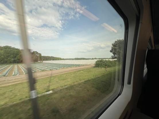 A wide angle picture of and through a Train window with blue sky, crop fields and a blurry pole outside