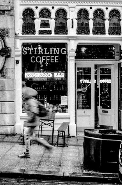 Black and white film photo of a coffee shop with a person rushing past. It's been raining all morning and the person has their coat hood on. The coffee shop has a neon sign saying 'Espresso Bar" underneath the store name "Stirling Coffee". Above the main window are small elaborate stain glass windows.