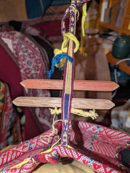 A narrow woven band has an X pattern in purple close to the camera. Two shed sticks are balanced in the weaving. A bundle of blue threads and a loop of yellow yarn are stationed in the unwoven warp threads. And at the far end, the patterned X and O in purple and yellow receed away to the anchor point.