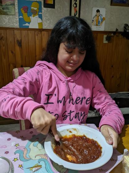 Una joven con cabello largo y oscuro está sentada a la mesa, vistiendo una sudadera rosa con la frase "I'm here to stay". Está mezclando algo en un plato blanco que parece ser una salsa o guiso de color rojo. La mesa tiene un mantel con dibujos de unicornios y estrellas. En la pared detrás de ella, hay varios cuadros, incluyendo uno de Homero Simpson sosteniendo una dona y otro de un jugador de fútbol con la camiseta de Argentina.