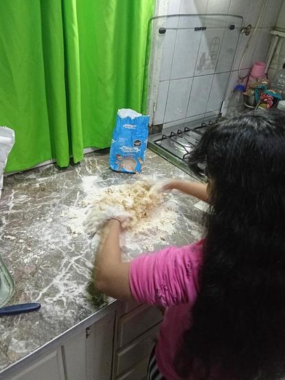 Una persona con cabello largo y oscuro está amasando una mezcla de harina sobre una encimera de mármol. Lleva puesta una camiseta rosa. En la encimera, hay un paquete de harina de la marca "Blancaflor" que está abierto. Al fondo, se ve una cortina verde y una cocina con una tapa de vidrio. También hay algunos utensilios de cocina y objetos en la esquina derecha de la imagen.