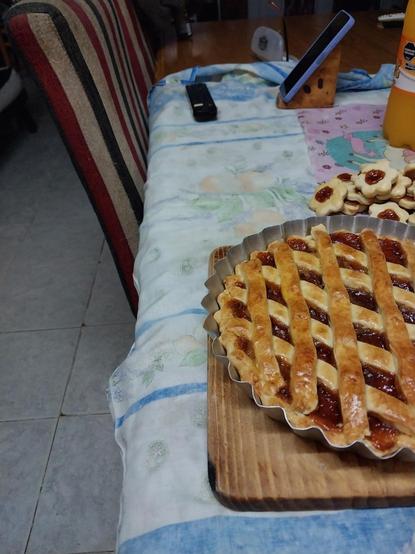 Sobre una mesa cubierta con un mantel de colores claros y estampado floral, hay una tarta con una rejilla de masa dorada y relleno de mermelada. Al lado de la tarta, hay galletas pequeñas con forma de flor, también con mermelada en el centro. En la esquina superior izquierda, hay una botella de jugo de naranja y un teléfono móvil. La silla junto a la mesa tiene un cojín con rayas de colores rojo, beige y negro.