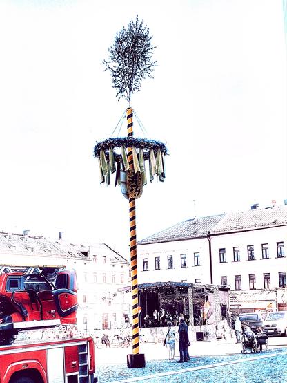 Farbenreduziertes Bild. In der Mitte der Maibaum. Links ein Teil eines Feuerwehrautos. Zwischen Maibaum und einer Häuserzeile eine überdachte Bühne mit Musikern.