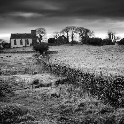 Black and white photo of a drystone wall, curving across rough pasture land, towards a church with a tower, trees and the gable end of a ruined church on the horizon.