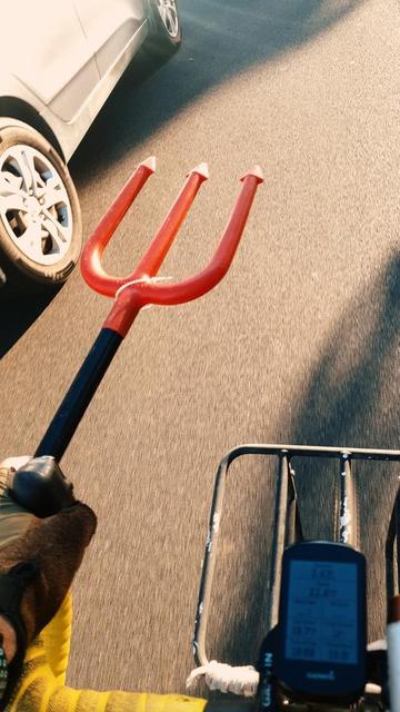 Bike rider's view of handlebars, left hand, and front wheel of a bike while riding. The left hand is gloved, and holding a plastic devil's trident, as made famous by Didi over the years in the TV coverage of Le Tour de France