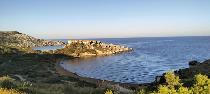 Looking from the top of the Ghajn Tuffieha trail down into the horseshoe of the sandy beach of Riviera Bay, the left side of the bay is protected by a rocky outcrop, and just beyond it another horseshoe-like bay is visible beyond the natural protection. The immediate foreground is bright green scrubland which contrasts against the calm deep blue of the sea, and light pink, pale blue if the horizon
