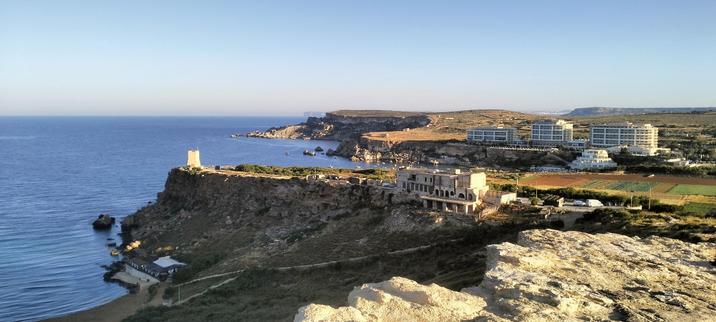 Rocky West coast of Malta at Ghajn Tuffieha (Riviera Bay), standing up on the hill looking down towards, the three Radisson owned hotels, that sit in on the outer limits of Majjistral nature park that rises into the distance. The island of Gozo is just visible through the haze or a pale blue horizon, that contrasts with the deeper blue of the Mediterranean sea.