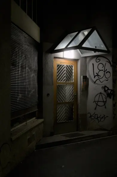 A residential building entrance doorway in a street by night lighted by a lamp above the door under the glass porch. The walls around the door are covered in graffiti.