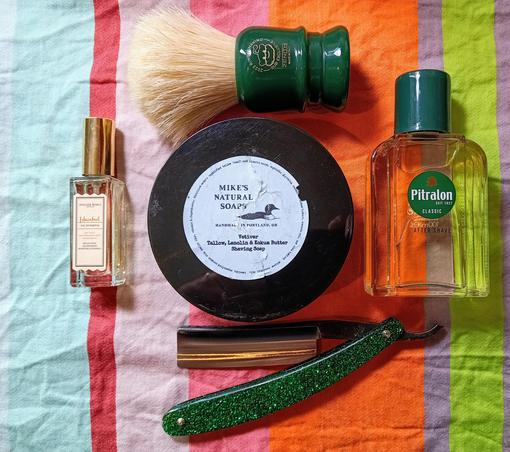 (Le Chat Mistral description)

This image shows a collection of grooming items arranged on a colourful striped cloth. There's a green shaving brush with a resin or plastic handle, a round container of Mike's Natural Soaps shaving soap, a green-handled straight razor, a small bottle of Atelier Rouge cologne, and a bottle of Pitralon aftershave. The items are neatly placed, suggesting a shaving routine setup. The colours are vibrant, with greens and oranges standing out against the striped background.
