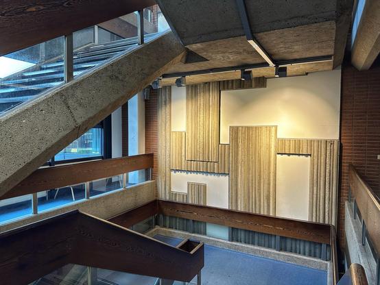 Ein architektonisches Interieur mit einer Treppe mit Beton- und Holzelementen, großen Fenstern, die natürliches Licht ermöglichen, und einer strukturierten Wand mit verschiedenen Paneeldesigns.