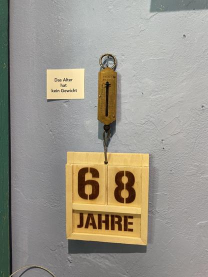A spring scale hangs on a nail on a grey wall. On the hook of the spring scale hangs a wooden sign saying '68 years'. To the left of the scales hangs a sign saying 'age has no weight'