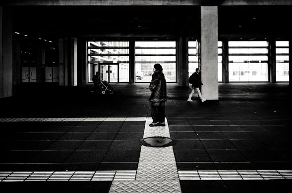 Within the monochromatic frame, several individuals traverse a public space. In the foreground, a figure clad in a fur coat stands upon a tactile paving surface, facing away from the viewer, close to a manhole. Further back, near the glass doors of a building, a person pushes a stroller. Another figure walks along the right side of the frame.