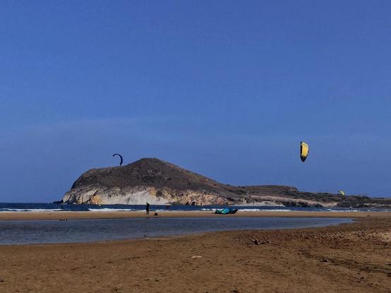 Vista de la playa de Genoveses. Al fondo se ve la duna petrificada. Hay varias cometas volando en el cielo y otra sobre la arena. Una persona está de pie, mirando al mar. En primer plano queda una pequeña laguna de agua de mar.