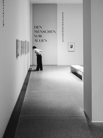 A woman leans in closely to examine framed artworks on a gallery wall, in a clean, minimalist museum space. Large text on the wall reads "DEN MENSCHEN VOR AUGEN" and a vertical inscription states "privat und offiziell, ideal und grotesk," enhancing the contemplative atmosphere of the exhibition.
