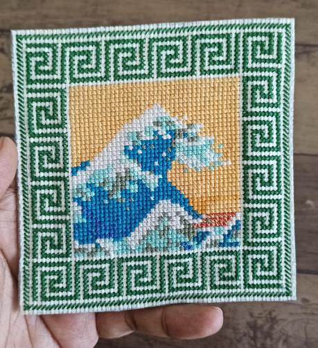 a rendition of the great wave off kanagawa,  with a yellow sky and blue,green, white threads to design the wave. 

there is a green border designed all around.