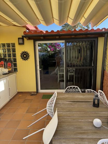 An outdoor patio features a wooden table surrounded by white chairs. A sliding glass door leads to the interior, with a decorative wall feature nearby. The space is shaded by a striped awning and is adorned with potted plants, including blooming flowers.
