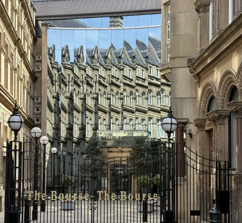 An old golden stone building reflected multiple times by the windows of a modern office. It’s sunny with a glimpse of blue sky.