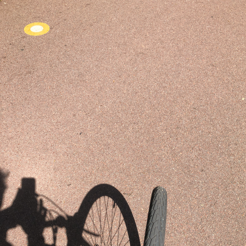 Minimalist photo of a pink-ish paved bike path. In the lower left of the frame  part of the front wheel of a bike and its shadow are visible. In the top left of the frame a yellow and white marking on the pavement can be seen.