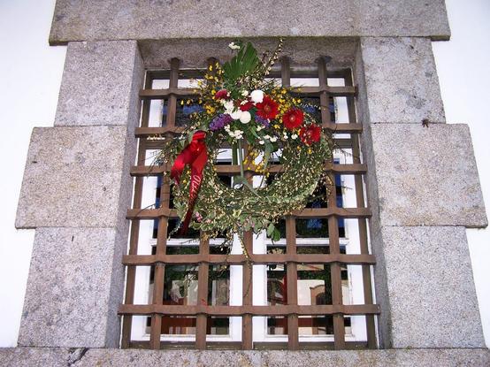 A imagem mostra uma coroa de flores pendurada no gradeamento de ferro forjado de uma janela oitocentista com uma moldura em pedra numa parede branca. A coroa é composta por uma variedade de flores coloridas, incluindo vermelhas, brancas e amarelas, com folhas verdes e um laço vermelho brilhante. A janela tem dois panos com seis vidraças e caixilharia de madeira pintada de branco. São visíveis os reflexos de janelas do outro lado da rua.