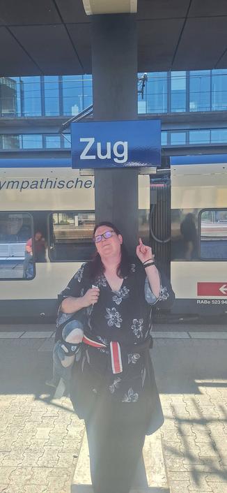 It me! Standing under the station sign for Zug. There's a Stadler FLIRT behind me in SBB white.
I'm wearing a light flowery top, coat tied at my waist & purple hair down