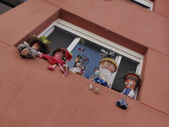 The orange-brown painted front of a house, photographed from below, diagonally in the middle of the photo (from top left to right) a window with white frames, recessed so that a border is created at the bottom. On the border 2 self-made dolls with hats and 3 plant pots decorated as dolls. The pots have legs and faces are painted on them. The legs of the rag dolls and the pots hang over the edge of the windowsill.