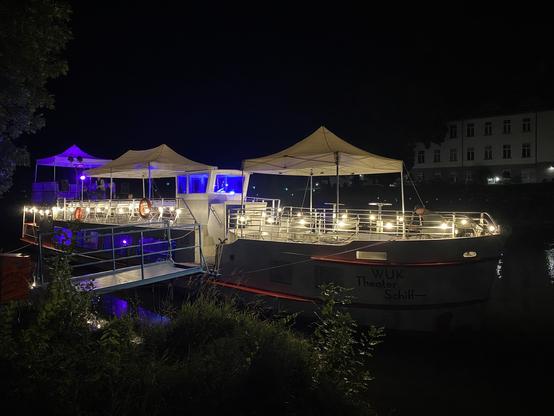 Ein hell erleuchteter Flussdampfer, am Ufer eines Flusses, bei Nacht - das WUK-Theater-Schiff.
Ein Steg führt links zum Schiff, auf dem drei weißer Pavillons stehen.
Links, am Heck, ist eine Bühne, die violett beleuchtet ist.
Rechts, am Bug, sitzt ein einsamer Mann und schaut auf sein Mobiltelefon.